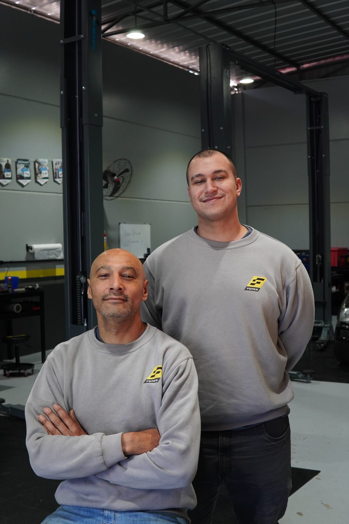 Dois mecânicos da equipe Feres posando lado a lado na oficina, uniforme cinza com logo Feres